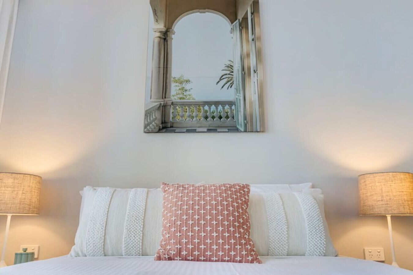 Ein Hotelzimmer mit einem Bett, zwei Nachttischlampen, einem Zier-Spiegel und Blick ins Freie.