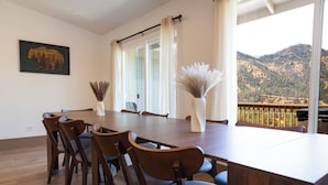 A dining area with a wooden table and chairs, a vase with dried plants, and a view of a mountainous landscape through large windows. - Shasta Lake Beautiful House w/ Hot Tub, BBQ, Ping Pong & Foosball Table (Redding)