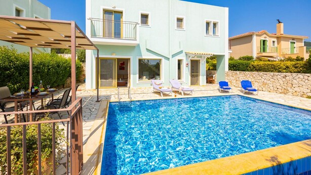 A swimming pool with lounge chairs, a covered patio, and a light blue building with balconies. - Villa Pinelopi With 2 Bedrooms and 3 Bathrooms (Kefalonia)