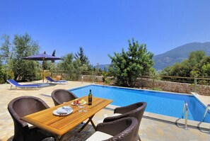 A poolside area with a wooden table, wicker chairs, and a blue umbrella. - Villa Irineos With 2 Bedrooms and 3 Bathrooms (Kefalonia)