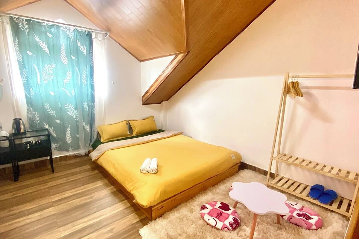 Un dormitorio pequeño y acogedor con cama, mesita de noche, estante de madera y ventana con cortina.