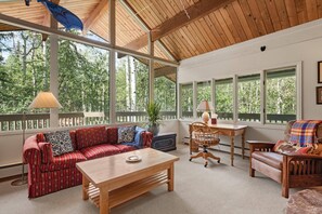 A spacious living room with a wooden ceiling, large windows offering a view of trees, a red striped sofa, a wooden coffee table, and a wooden chair. - Unique Mountain Home! Spacious Deck & Hot Tub, Wood Stoves, Balconies with Tranquil Forest Views (Aspen)