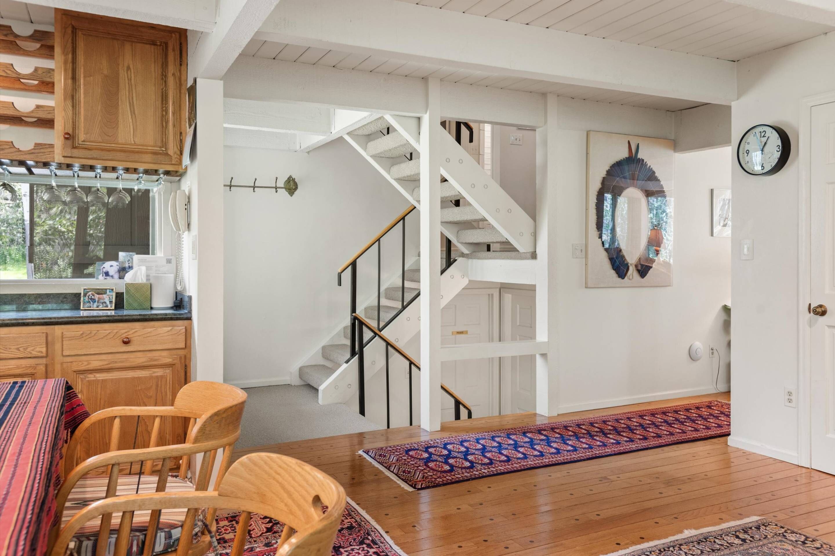 A modern kitchen with wooden cabinets, a dining area with a patterned rug, and a staircase leading to another level.