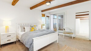 Un dormitorio con cama blanca, mesita de noche, silla y ventana grande con vistas.