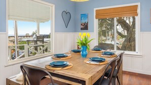 Un comedor con mesa de madera dispuesta para seis personas, un jarrón azul con flores amarillas, y vistas al exterior a través de ventanas amplias.