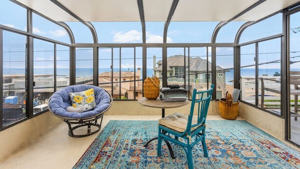 Un balcón con vista al mar, una silla azul acolchada, una mesa con cojines y una alfombra estampada.