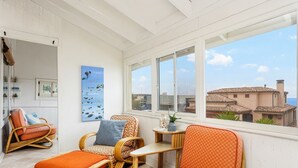 Una habitación luminosa con muebles de mimbre, un cuadro y vistas a edificios a través de grandes ventanales.