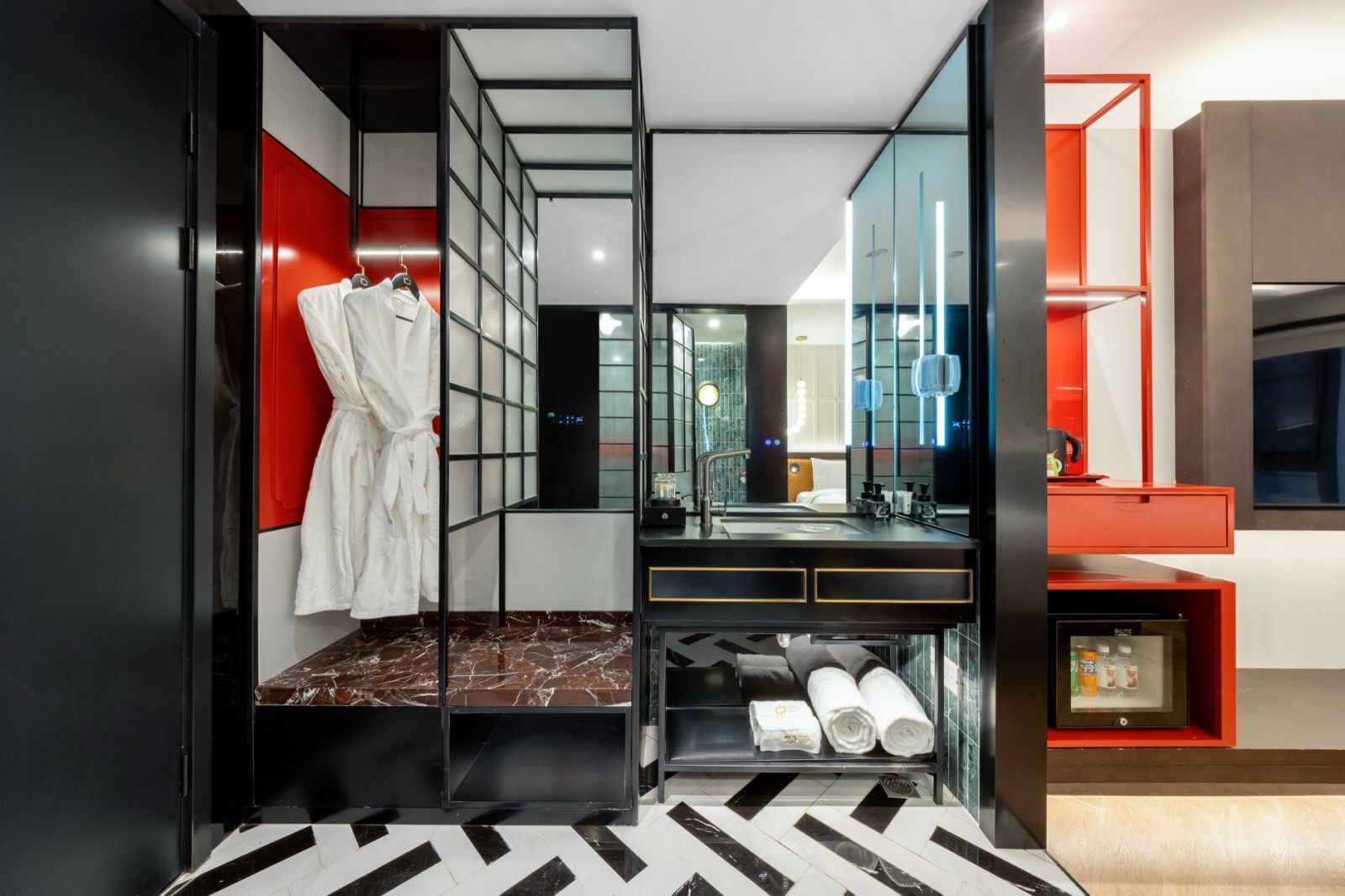 Un baño moderno con un espejo grande, piso de baldosas blanco y negro, separador de vidrio y una pared de acento roja.