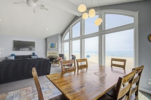 A spacious living area with a large wooden dining table, comfortable seating, a fireplace, and a view of the beach.