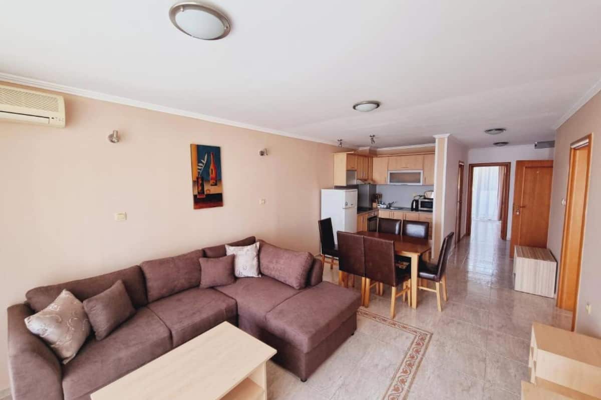 A living room with a sectional sofa, a dining table, and chairs. There is a kitchen area with cabinets and appliances visible in the background.