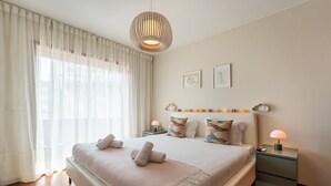 Ett modernt hotellrum med en stor säng, ett nattduksbord med en lampa och ett fönster med tunna, genomskinliga gardiner.