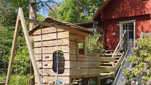 Puuinen leikkimökki, jossa on liukumäki ja kiipeilyseinä, ja sen ympärillä on nurmikkoa ja puita.