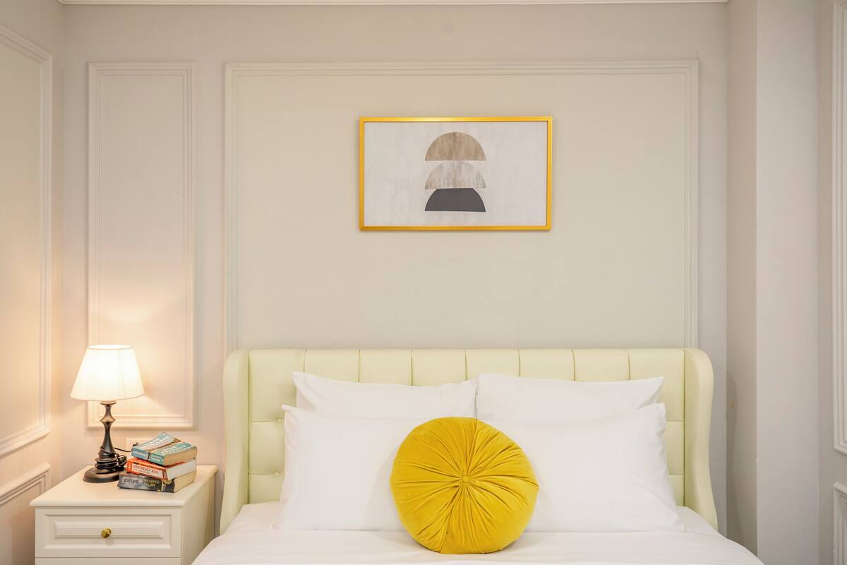 A neatly arranged bedroom with a bed, pillows, a bedside table with a lamp, and a framed artwork on the wall.
