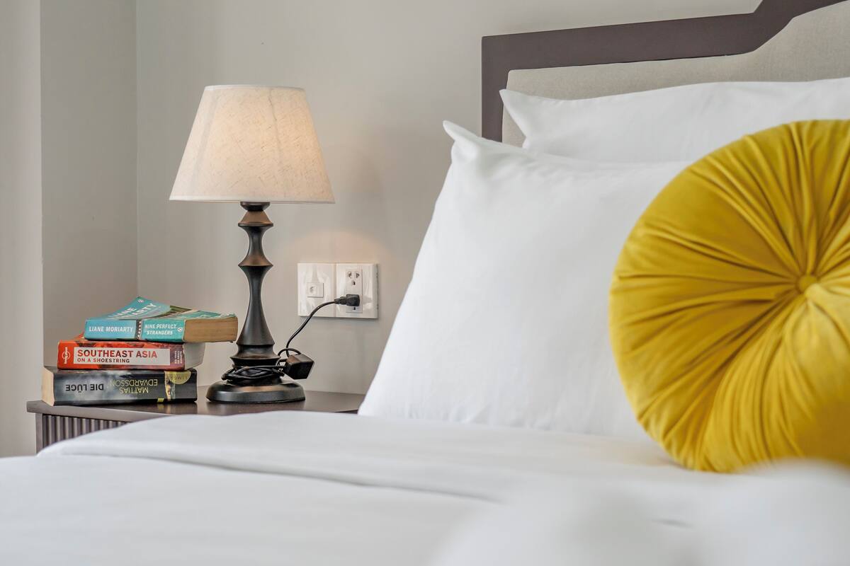 A neatly made bed with white linens and a single yellow accent pillow. A bedside table holds a lamp, a few books, and a power outlet.