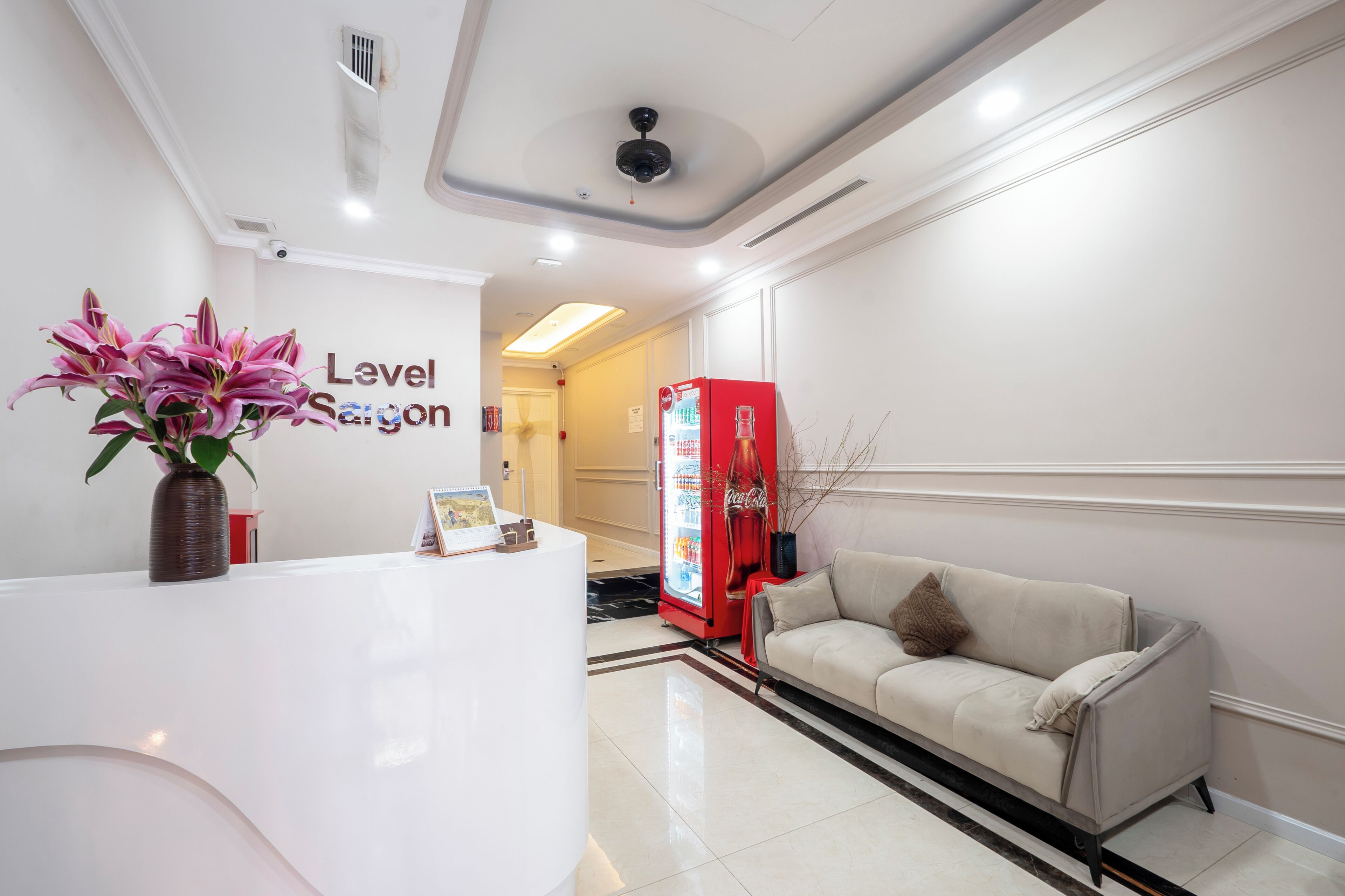 Uma área de recepção com balcão branco, um refrigerador vermelho da Coca-Cola, um sofá bege e um vaso com flores cor-de-rosa.