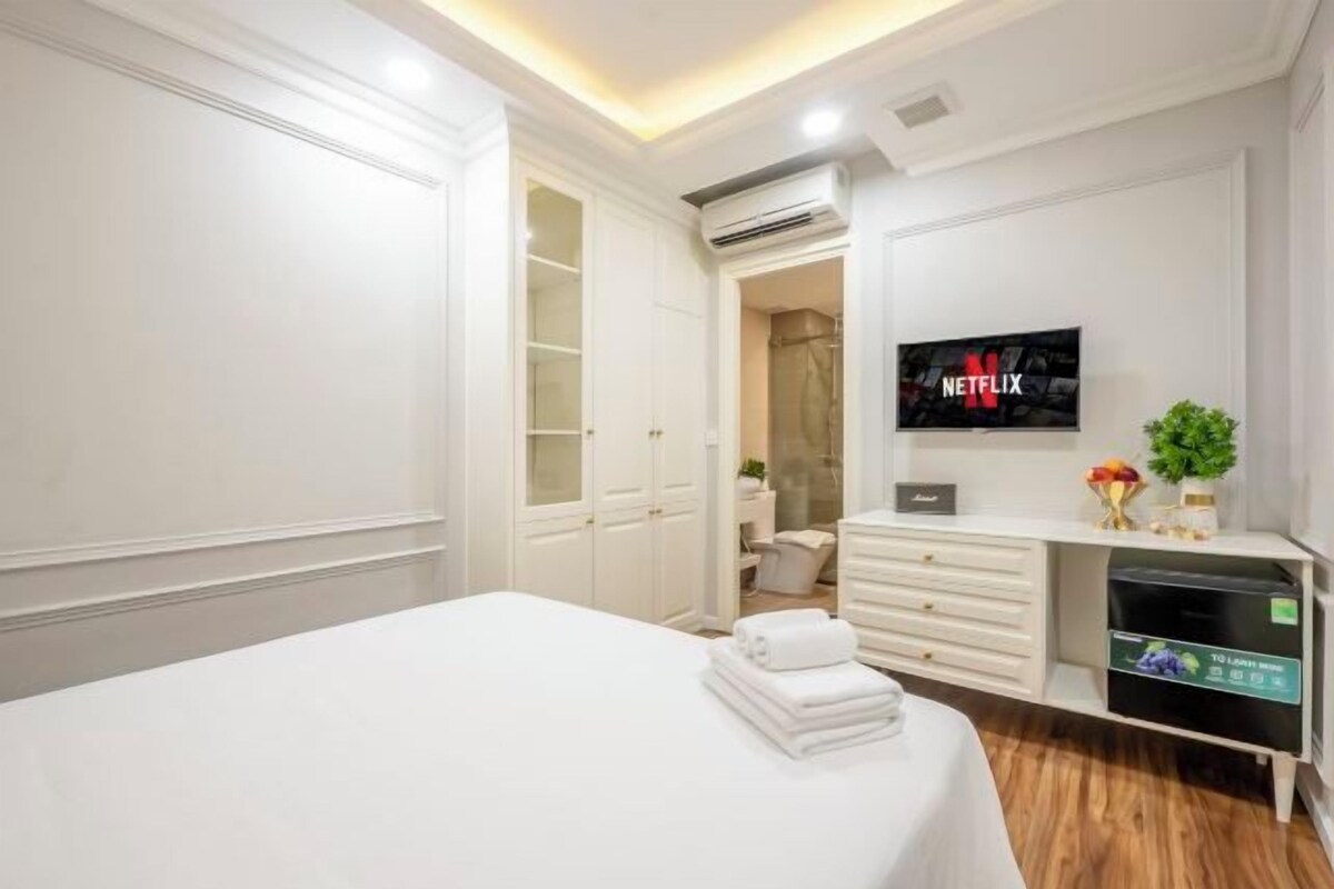 A hotel room with a bed, a TV mounted on the wall, a white wardrobe, and a small desk with a plant.
