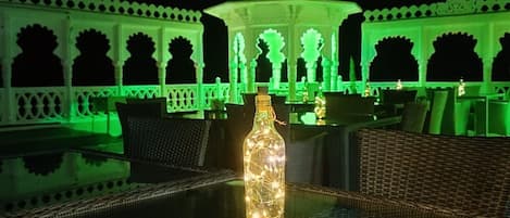 An outdoor seating area with a table and chairs, a bottle of lighted candles, and a building with arches and a dome in the background.