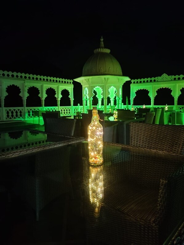 An outdoor seating area with a table and chairs, a bottle of lighted candles, and a building with arches and a dome in the background. - Arryaa Inn (Udaipur)