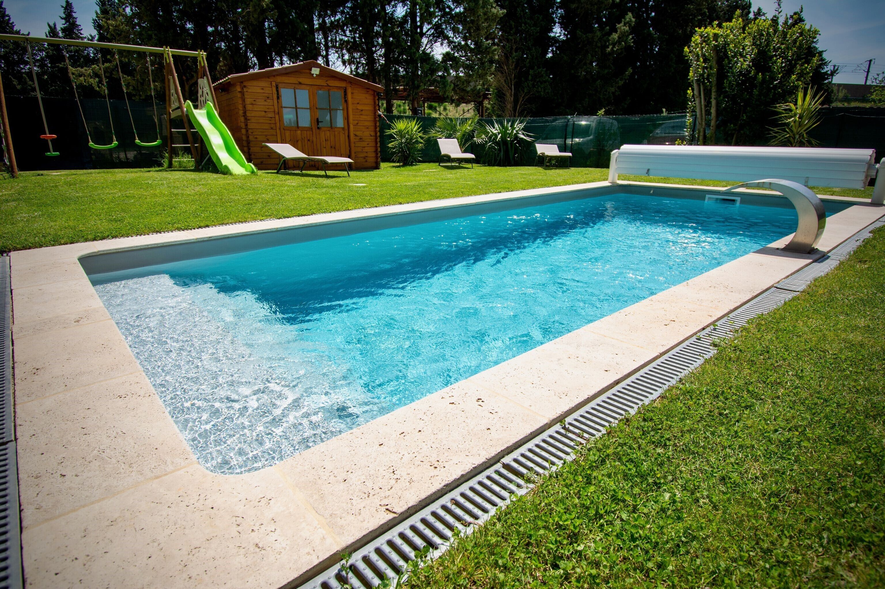 Una piscina rectangular amb tobogan integrat, envoltada per un prat de gespa i una cabana de fusta.