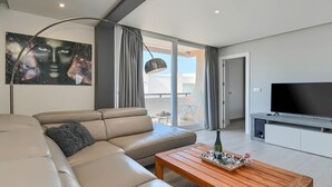 A modern living room with a beige sofa, a wooden coffee table, a flat-screen TV, and a large window with curtains.