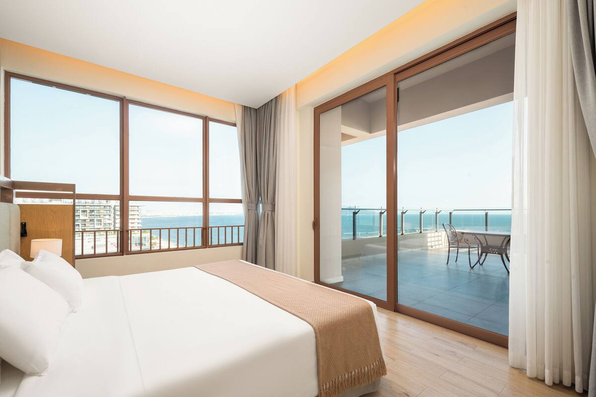 Habitació d'hotel amb un llit gran, un balcó amb vista al mar i una zona de menjador amb mobles d'exterior.