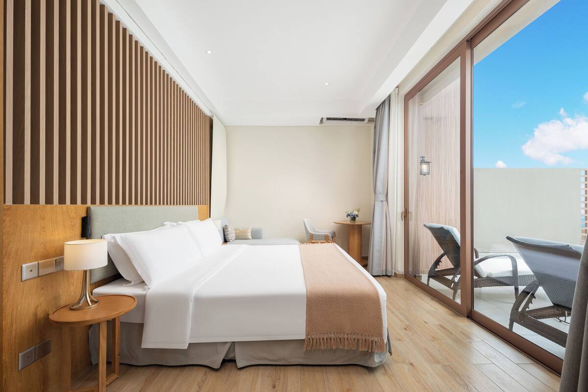 Habitació d'hotel moderna amb un llit gran, una taula de nit amb una làmpada, una taula rodona petita i una cadira. Hi ha una porta corredissa de vidre que dona a un balcó amb mobiliari exterior.