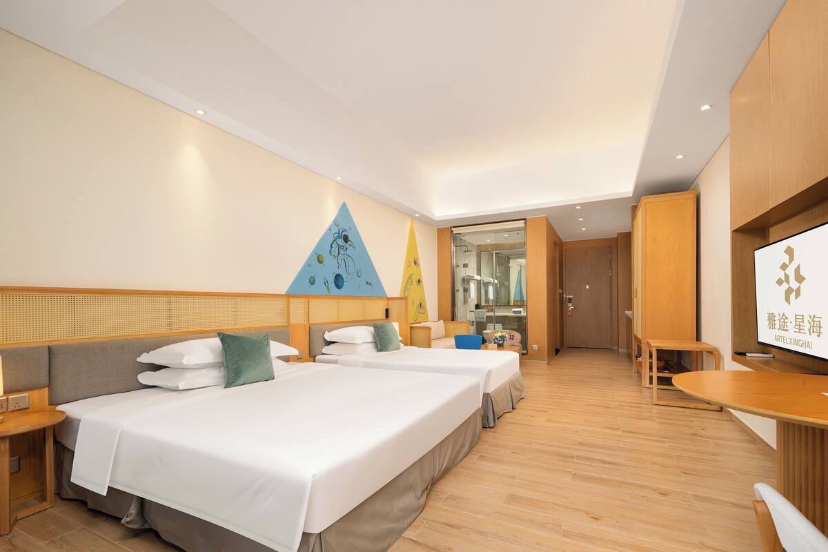 Habitació d'hotel amb tres llits, sòl de fusta, televisió i una porta de vidre que dona a un bany.