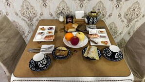 A dining table set with various dishes, including bread, fruits, and condiments, in a room with patterned wallpaper and a cushioned chair.