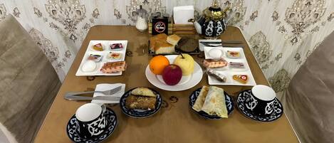 A dining table set with various dishes, including bread, fruits, and condiments, in a room with patterned wallpaper and a cushioned chair.