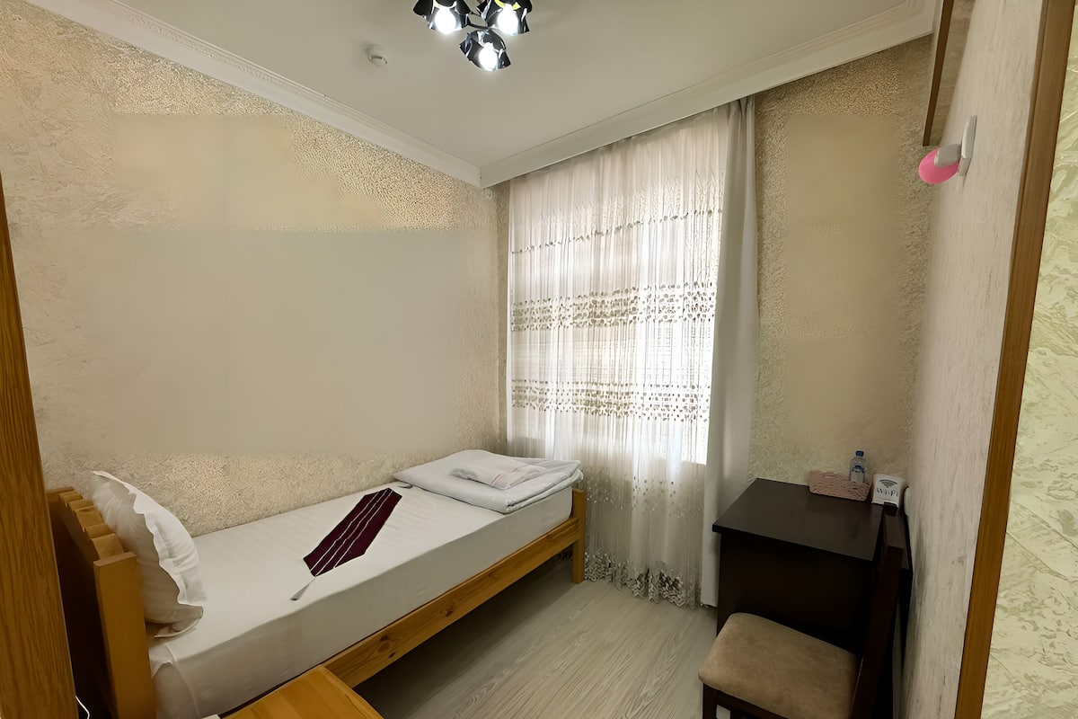 Una camera da letto piccola e ordinata con un letto singolo, una scrivania in legno, una sedia e un comodino con una lampada.