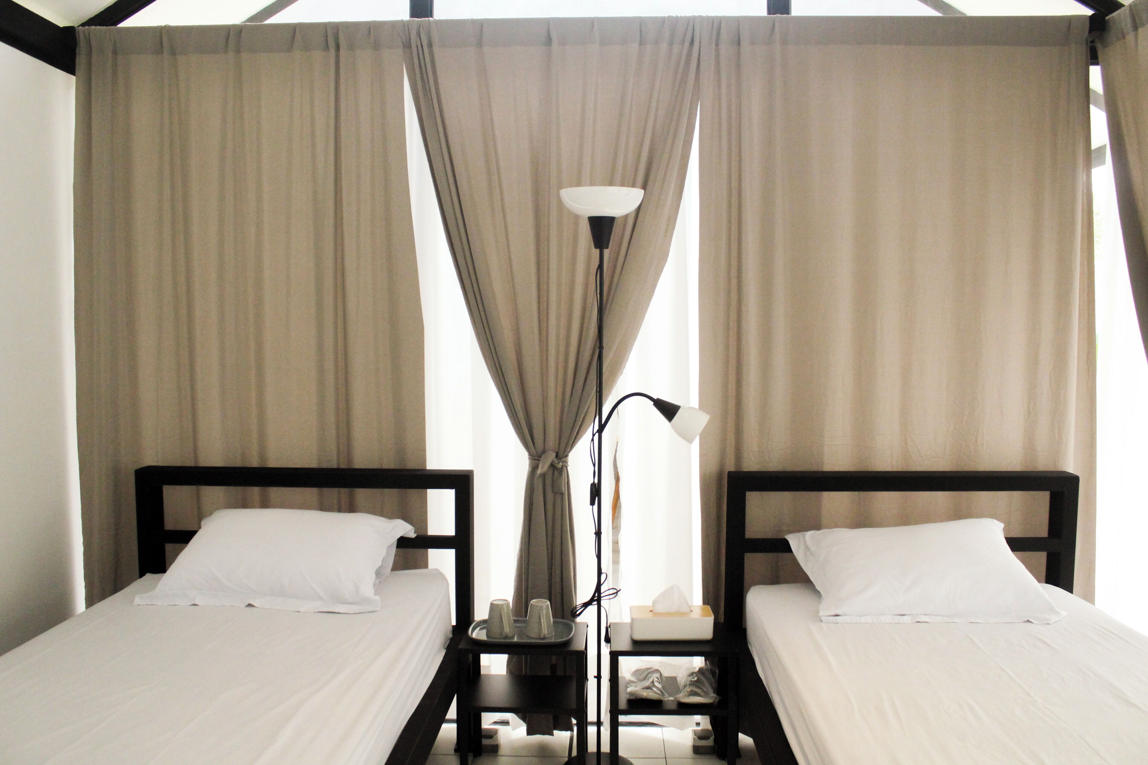 A hotel room with two single beds, a nightstand with a lamp, and a window with curtains.