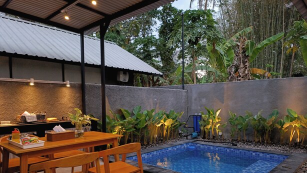 Un área de piscina con terraza de baldosas, sillas de madera, una mesa con aderezos y plantas a lo largo del borde de la piscina.