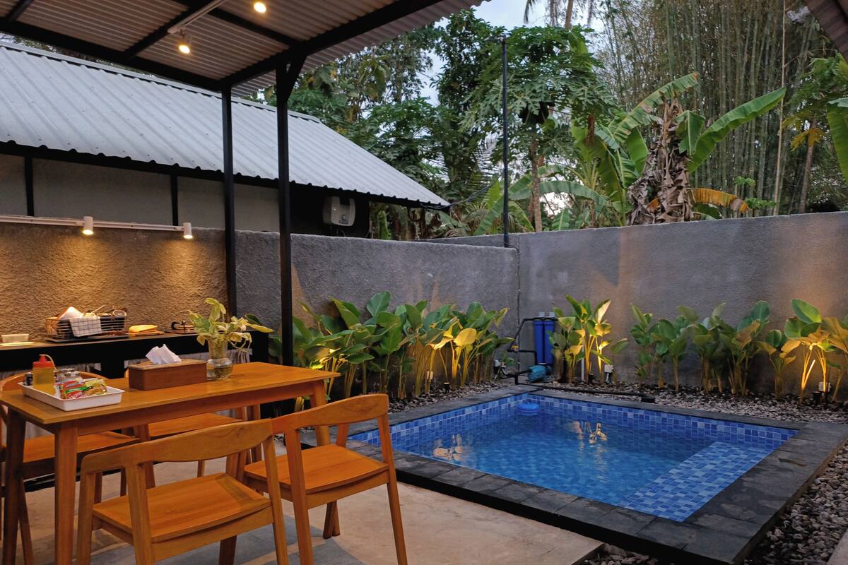 A pool area with a tiled deck, wooden chairs, a table with condiments, and plants along the pool's edge.