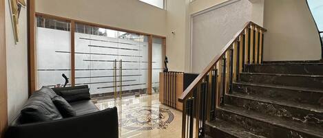 A spacious living area with a black leather sofa, a wooden reception desk, and a staircase with a wooden railing.