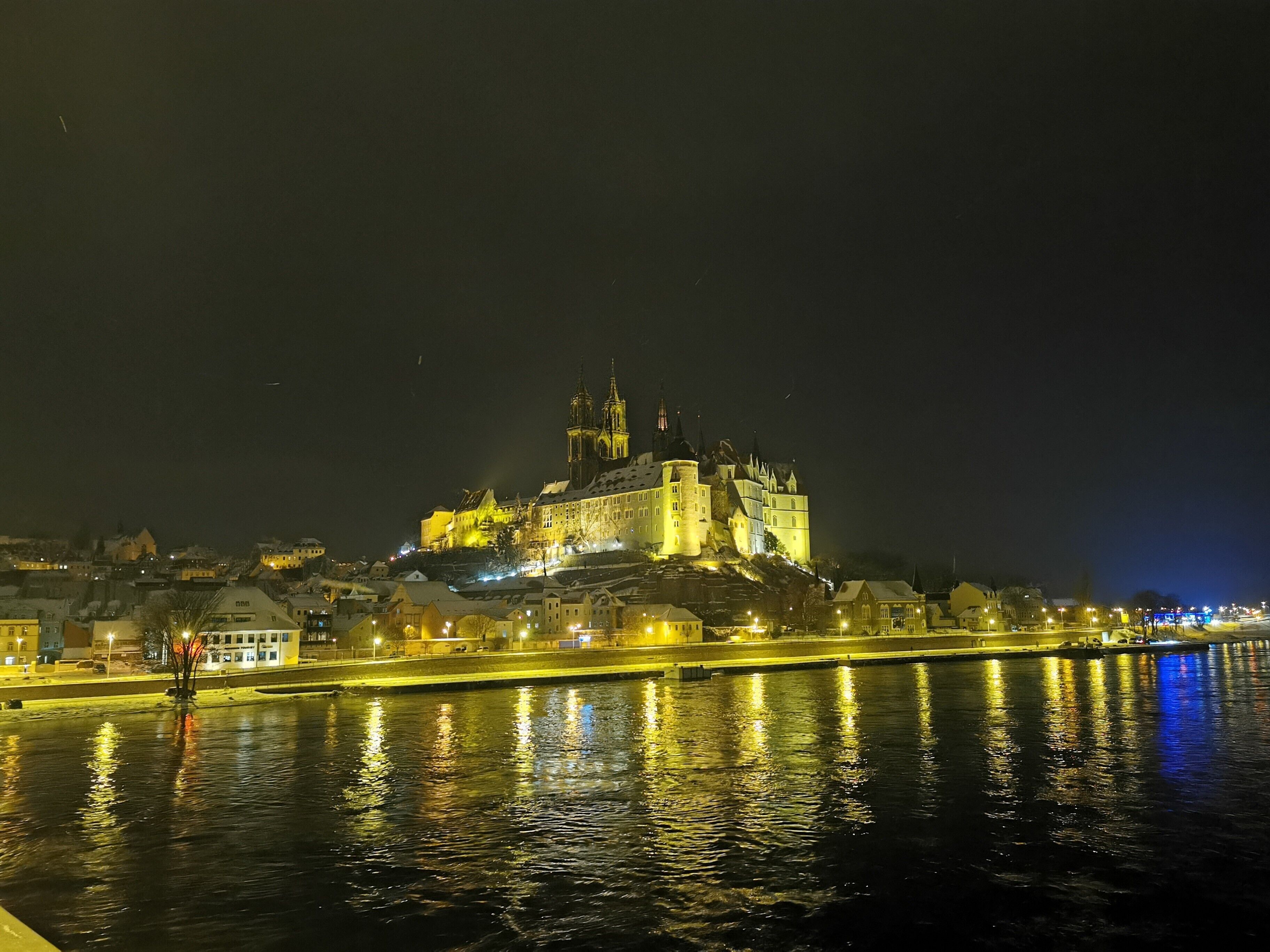 Eine schlossähnliche Struktur mit einem Burggraben, umgeben von Gebäuden und einem Fluss, der Lichter widerspiegelt.