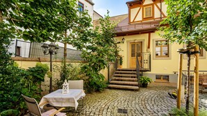 Une cour avec un chemin pavé de pierres, une table entourée de chaises et un escalier menant à un bâtiment à la façade en bois.