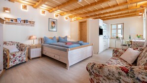 Une pièce chaleureuse avec un plafond en bois, un lit avec une literie bleue, un canapé à motifs floraux, une petite table avec une lampe, et un coin cuisine à l’arrière-plan.