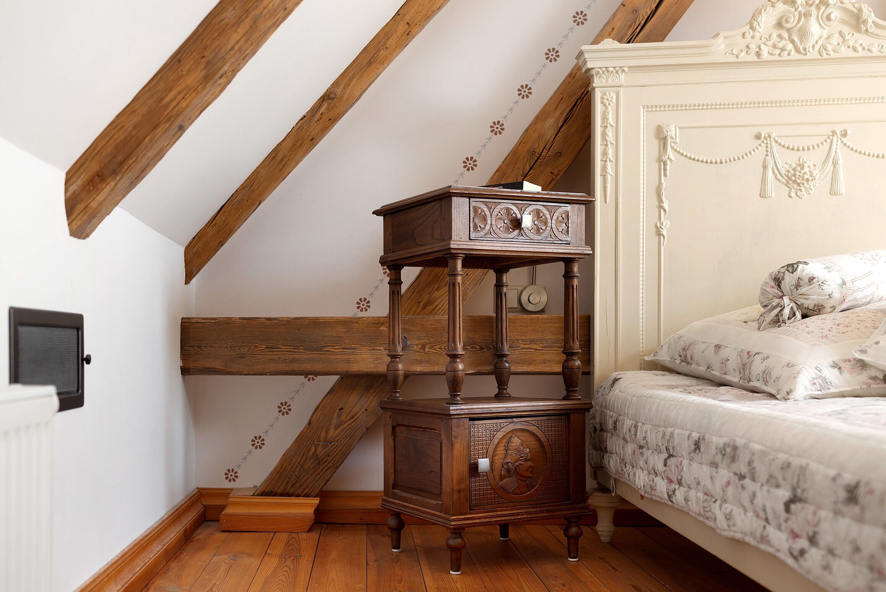 Una camera da letto con un letto in legno, un comodino, un termosifone e un soffitto spiovente con travi in legno.