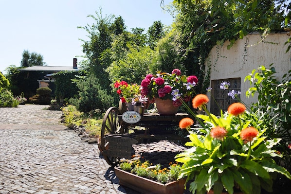 A cobblestone path leading to a rustic cottage with a garden cart filled with flowers and a sign welcoming visitors to the cottage.