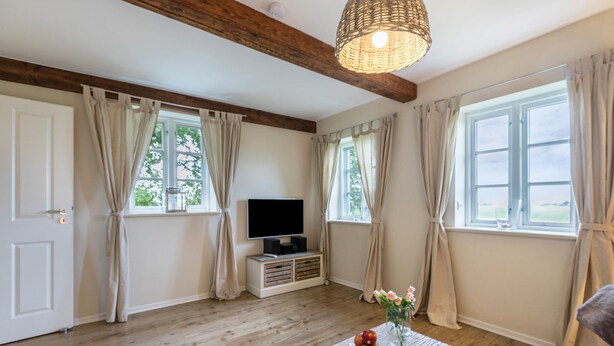 A room with wooden flooring, a flat-screen TV on a stand, a table with a book and fruit, and a window with curtains.