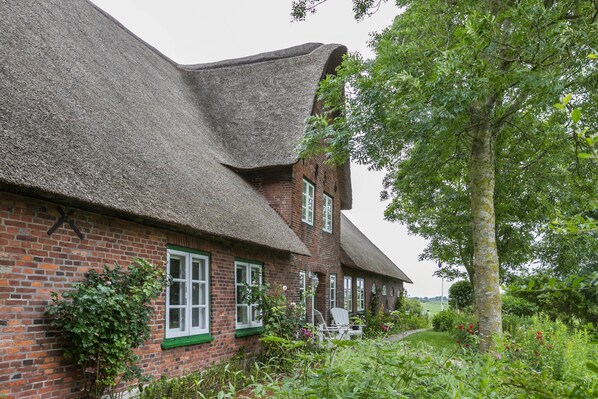 Une maison traditionnelle en briques, avec un toit de chaume, un jardin agrémenté d’un sentier et un arbre.