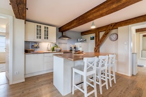 Une cuisine avec des meubles blancs, un îlot central en bois doté de tabourets, une horloge au mur et des poutres apparentes.