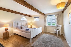 Une chambre avec un plancher en bois, un lit blanc, deux lampes de chevet, une fenêtre avec des rideaux et une chaise.