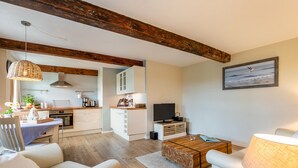 Una sala moderna con sofá, mesa de centro y lámpara; una cocina con armarios y televisor; vigas de madera en el techo; y un cuadro en la pared.