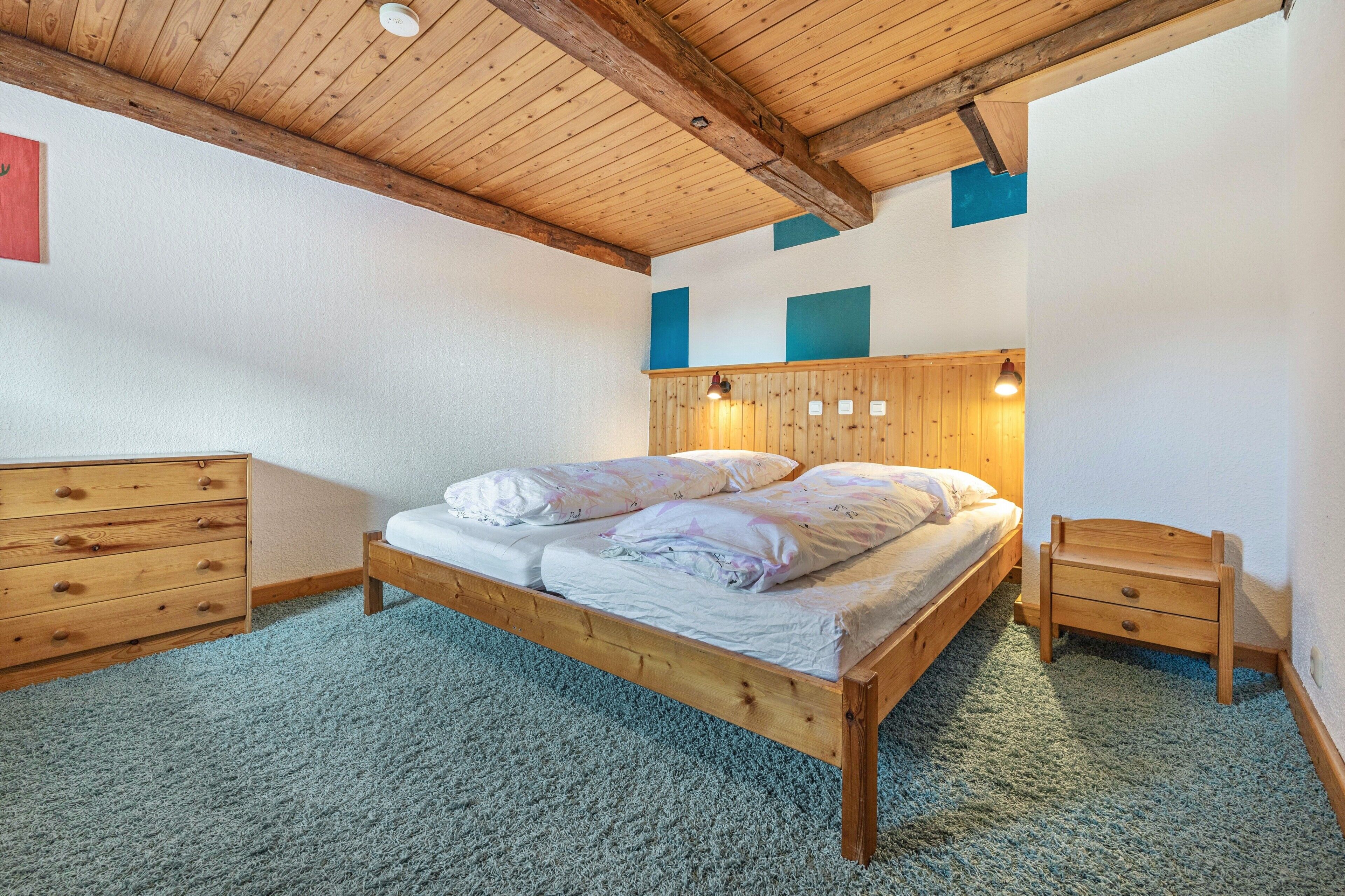 Quarto com cama de madeira, mesa de cabeceira, cômoda e teto com vigas aparentes.