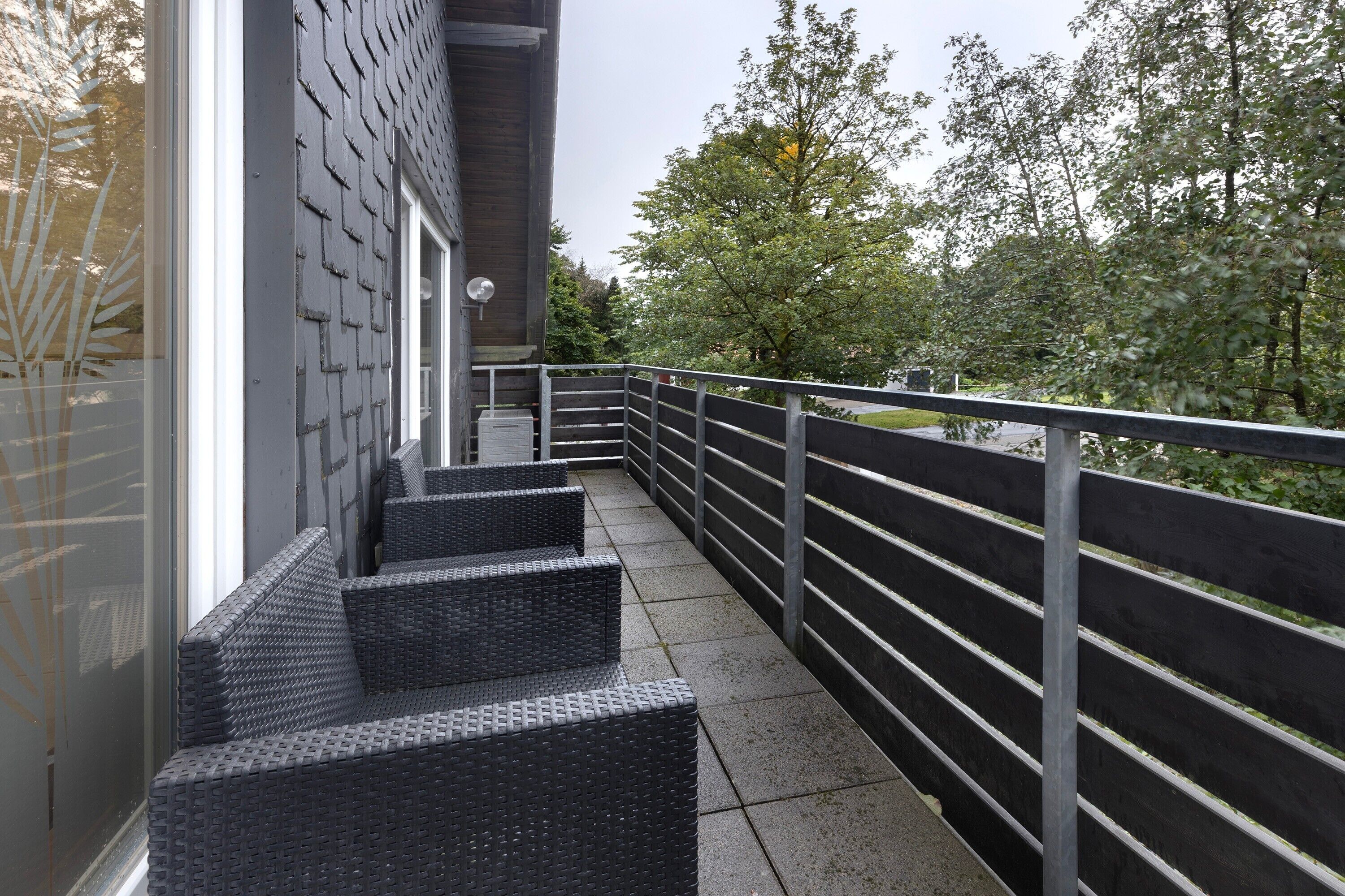 En balkon med fletningsmøbler, gelænder i metal og udsigt til træer og en bygning.