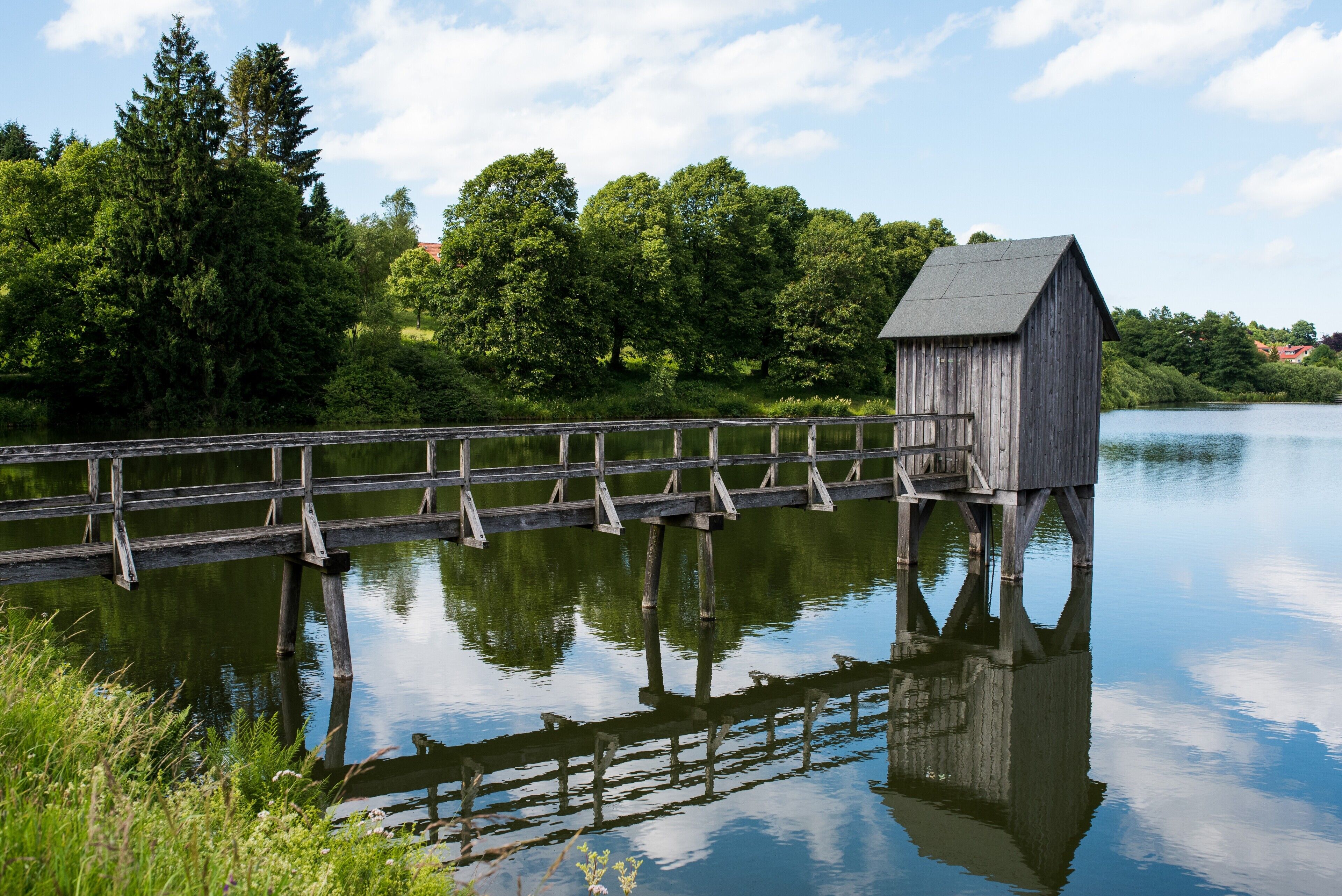 A wooden boathouse on stilts by a calm lake, surrounded by lush greenery and trees.