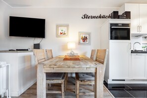Un coin repas avec une table en bois, des chaises, un téléviseur, des tableaux encadrés, et un espace cuisine avec des meubles blancs et des appareils électroménagers.
