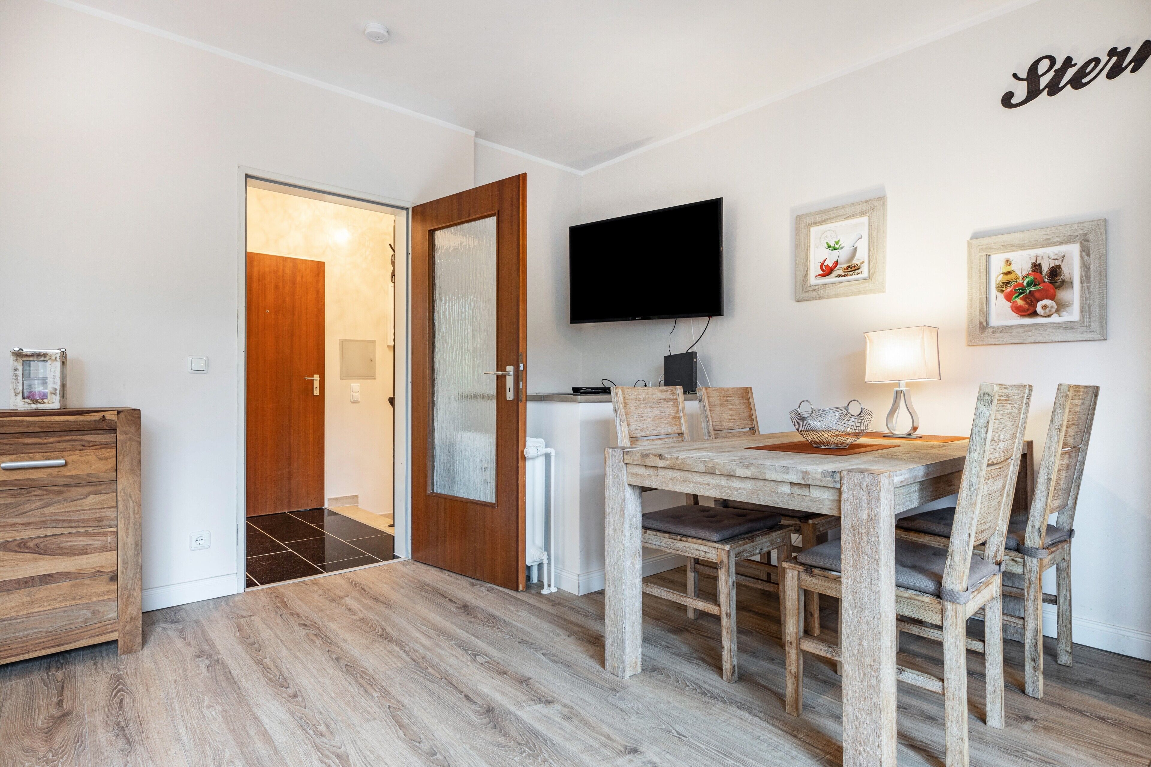 A dining area with a wooden table and chairs, a television mounted on the wall, and a door leading to another room.