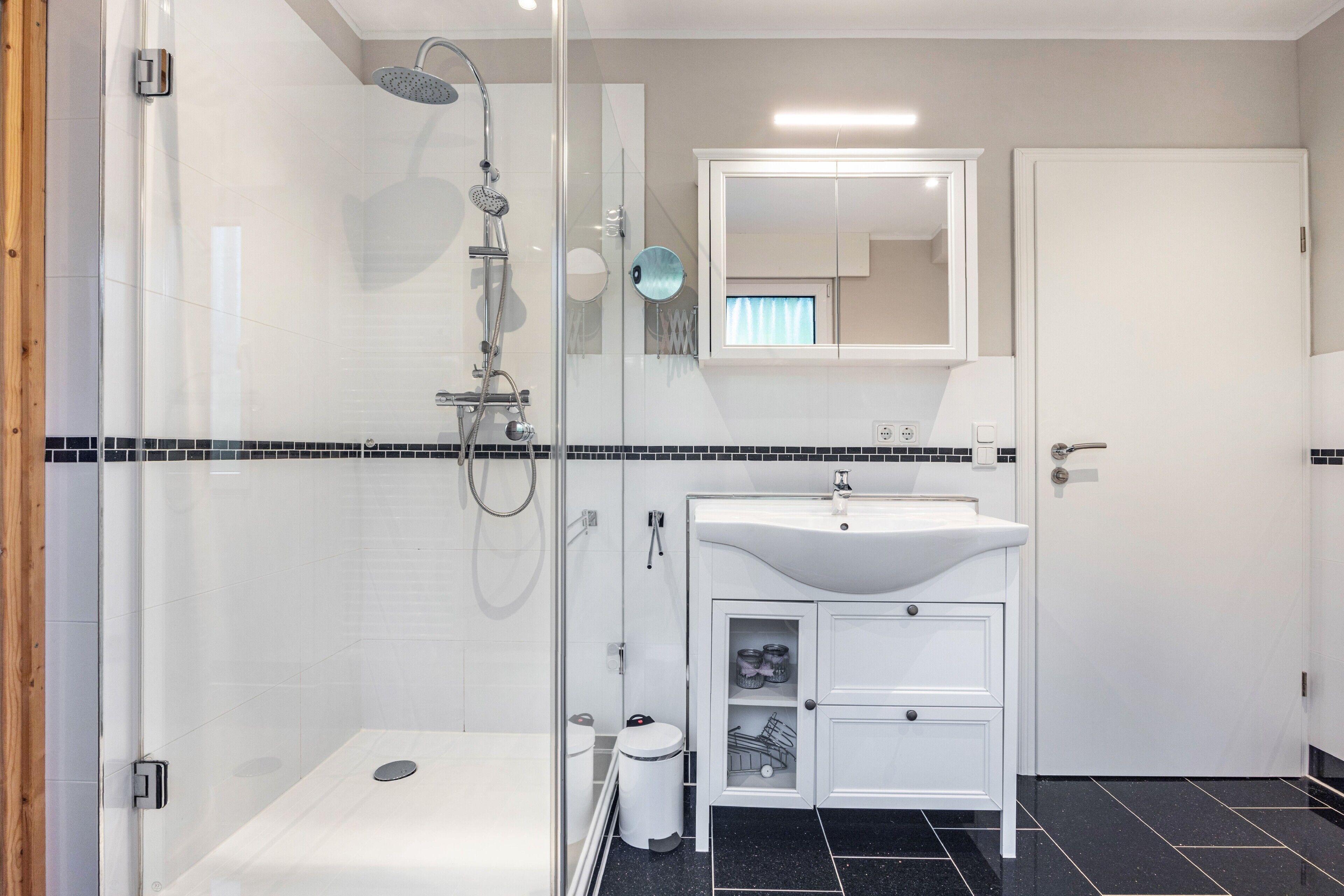 A modern bathroom with a glass-enclosed shower, a white sink with a mirror above, and a door to the right.